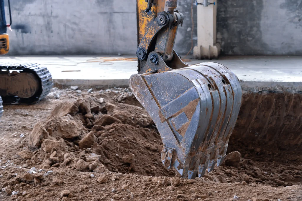 Excavator bucket performing ground disturbance work, highlighting the importance of permits and utility locating before digging.